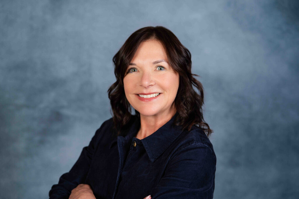 A professional headshot portrait of a woman with dark wavy hair smiling at the camera against a muted blue background.