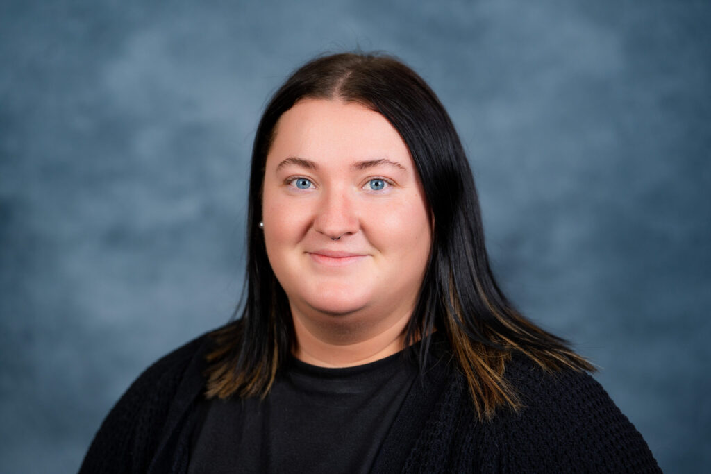 A professional headshot of a woman with long dark hair smiling at the camera against a neutral blue-grey studio background.