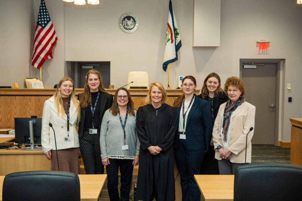 A professional group of nine people in business attire pose together in a formal courtroom or government building setting.