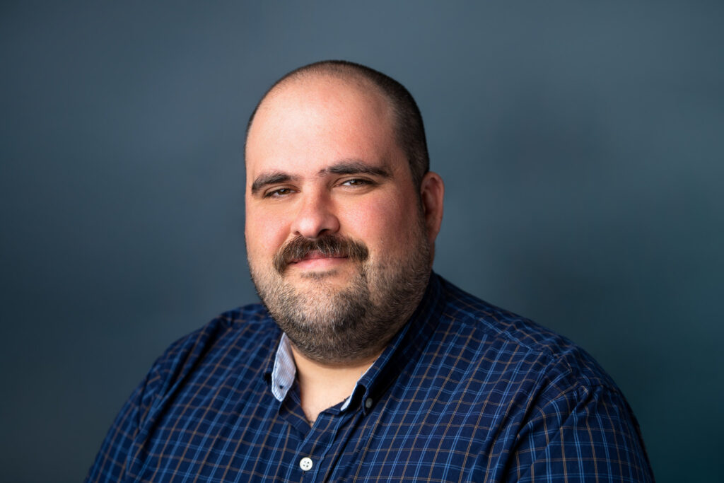 A professional headshot of a man with a beard wearing a blue plaid shirt against a neutral slate blue background.