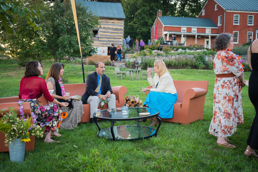 A small group of people participate in an outdoor garden workshop on a grassy lawn in front of a red brick building, with potted plants and wooden seating arranged in a casual circle.