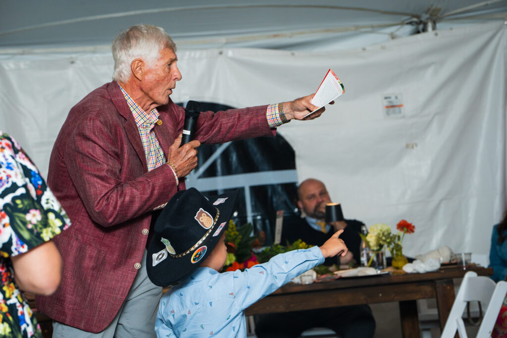 An auctioneer in a maroon blazer gestures expressively while speaking at a livestock or agricultural auction under a tent.