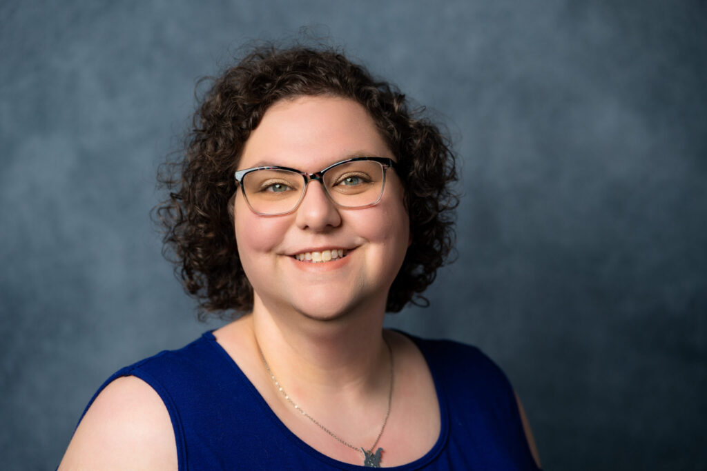 A professional headshot of a smiling person with curly hair and glasses wearing a dark blue sleeveless top against a textured gray-blue background.