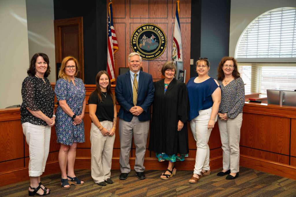 A formal group portrait of eight people posing together in an official government or civic venue with wood paneling, an American flag, and an official seal visible in the background.