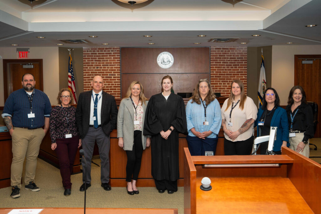 A professional group of approximately twelve people pose together in an indoor institutional setting with a wooden podium, brick walls, and formal lighting.