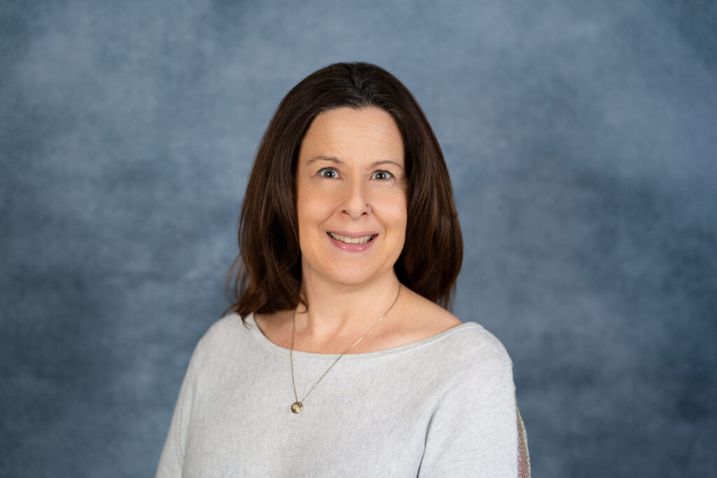 A professional headshot portrait of a smiling woman with dark hair wearing a light gray top against a neutral blue-gray background.