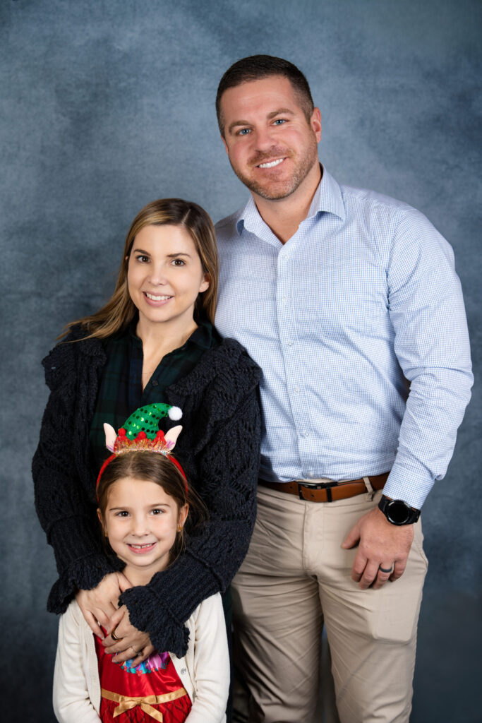 A professional family portrait of three people—an adult male, adult female, and young child—posed against a neutral blue-gray background.