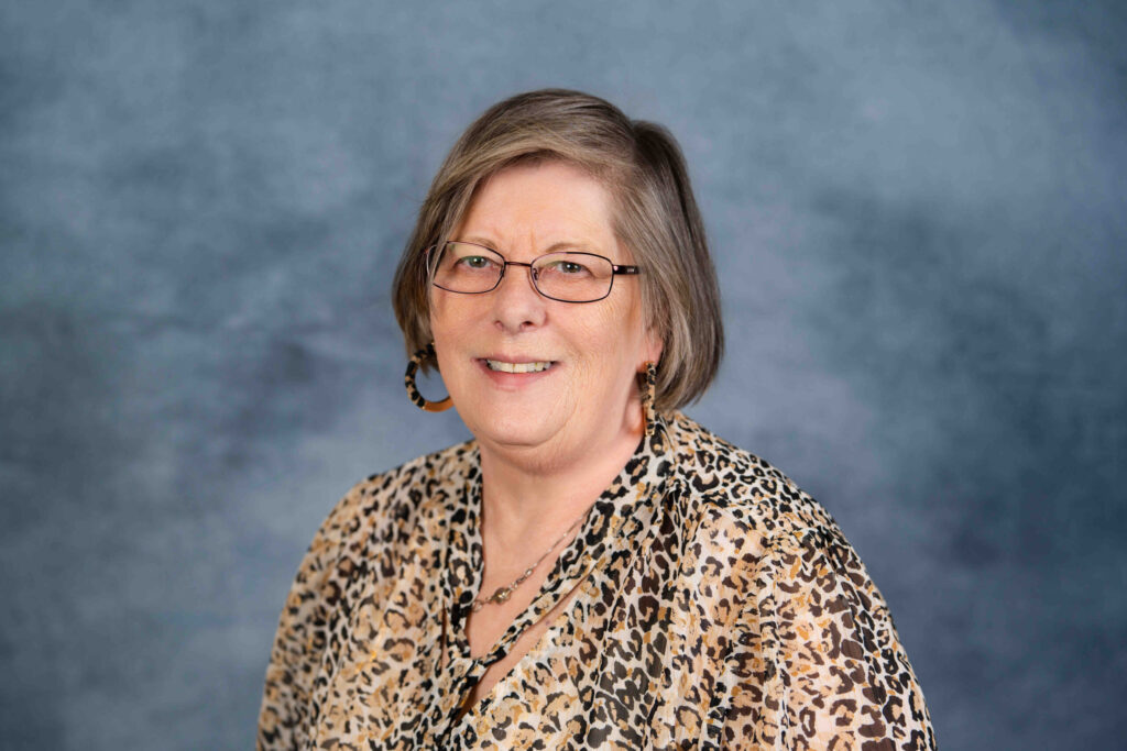 A professional headshot portrait of a middle-aged woman wearing glasses and a patterned blouse against a blue-gray background.