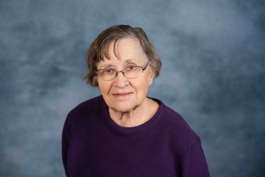 A professional headshot portrait of a woman wearing glasses and a purple sweater against a blue-gray studio backdrop.