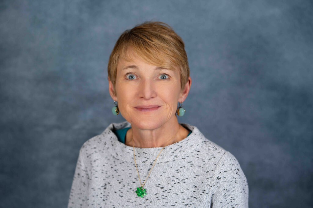 A professional headshot portrait of a smiling woman with blonde hair wearing a grey speckled sweater and green brooch against a blue-grey background.
