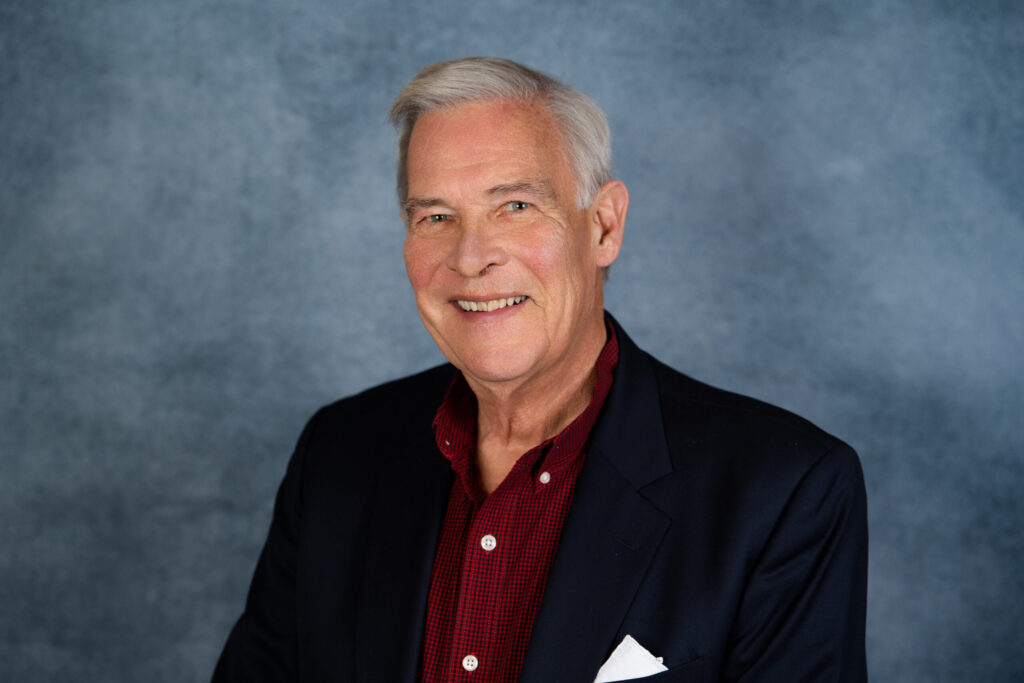 A professional portrait of a smiling older man wearing a dark blazer and burgundy shirt against a neutral gray background.