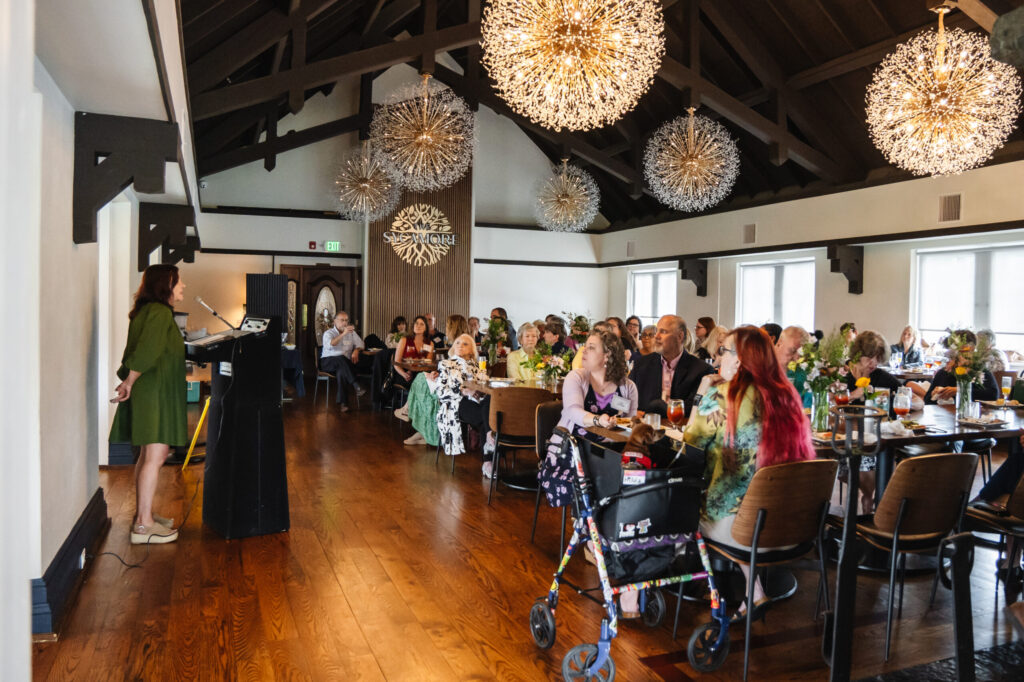 A lively indoor event or gathering taking place in a modern venue with exposed ceiling, decorative pendant lights, and wooden flooring, with numerous guests mingling and socializing.