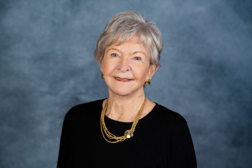 A professional portrait of a smiling mature woman wearing a black top and gold necklace against a textured blue-gray background.