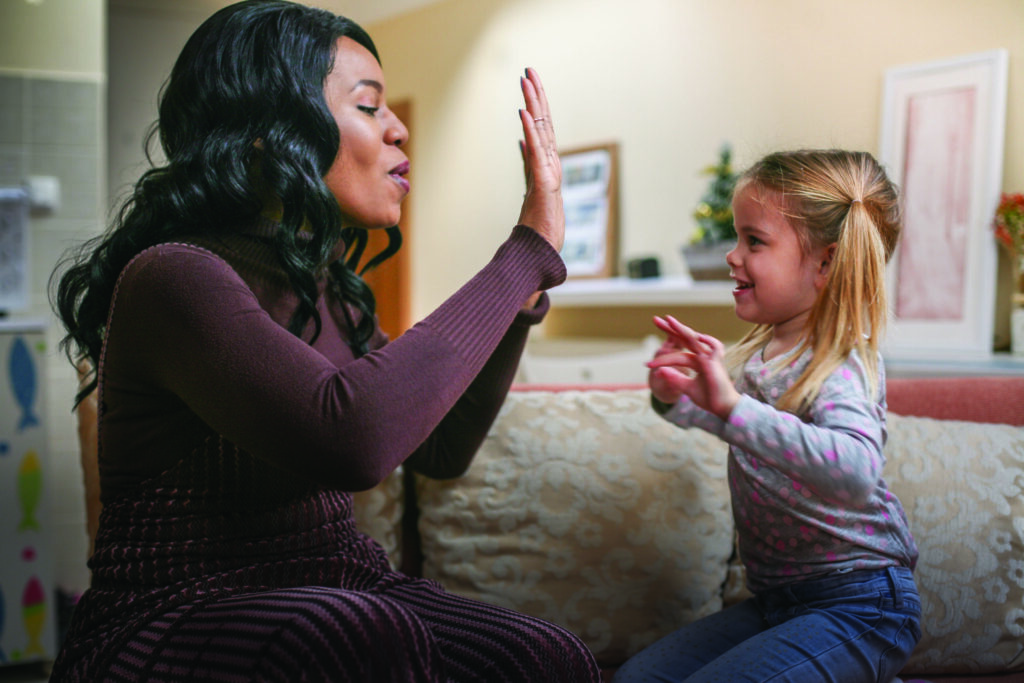 A woman and young girl engage in a playful hand-clapping game or interaction while sitting on a couch in a home setting.