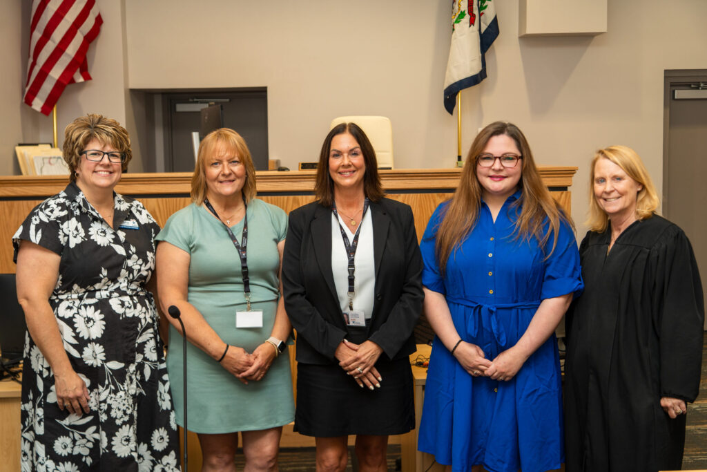Five professional women pose together in a formal indoor setting with wooden paneling and an American flag visible in the background.