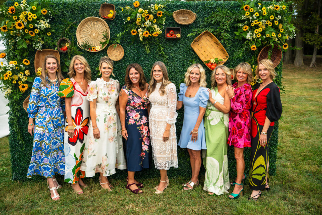 A group of women in colorful dresses pose together outdoors against a decorative floral backdrop with woven baskets and greenery.