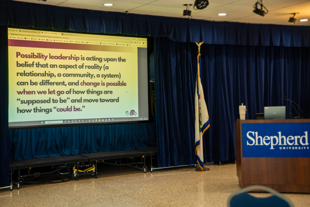 An indoor presentation setup with a projection screen displaying text, a podium, blue curtains, and Shepherd branding visible in an auditorium or conference room.