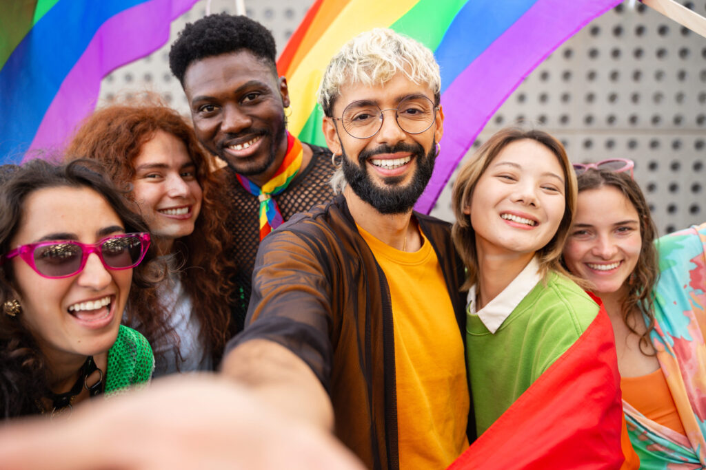 A diverse group of six young people take a selfie together in front of a vibrant, colorful rainbow-striped background wall.