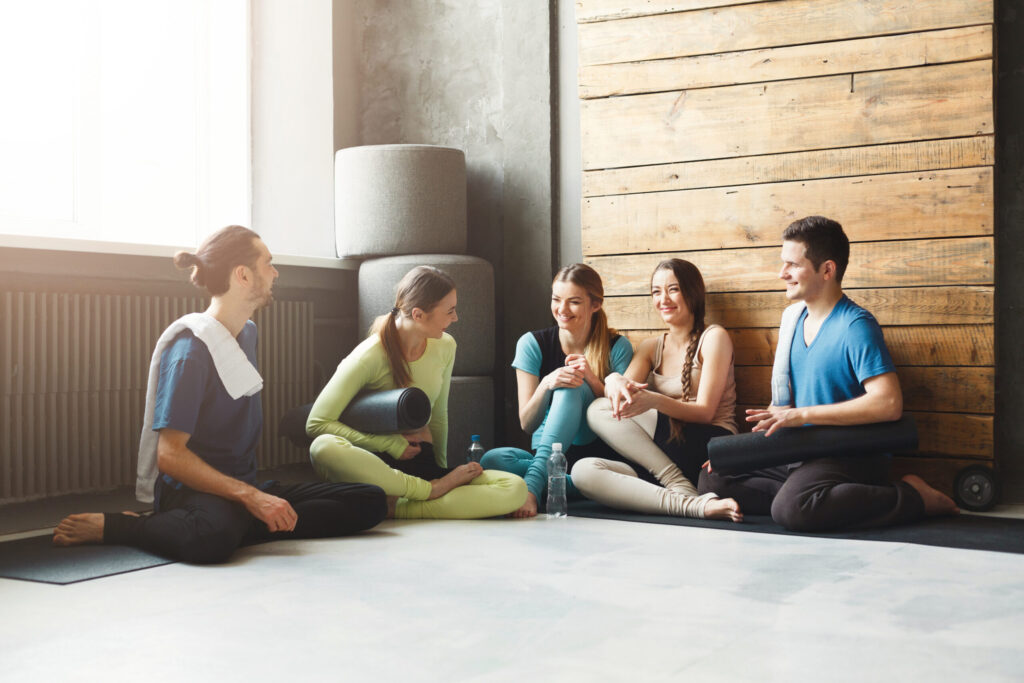 A small group of five people sit on yoga mats in a bright, modern fitness studio with wooden accent walls during a workout or wellness class.