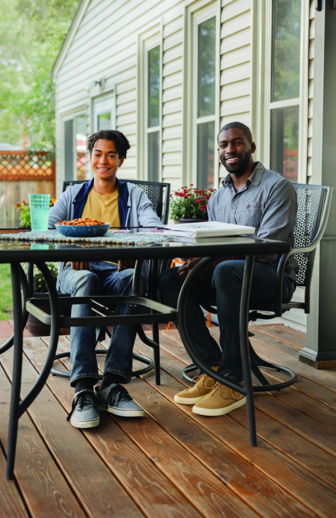 Two people sit at a table on a wooden deck outside a house, engaged in casual conversation.