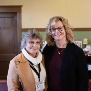 Two smiling women stand together for a formal portrait inside an indoor venue with warm beige and green walls.