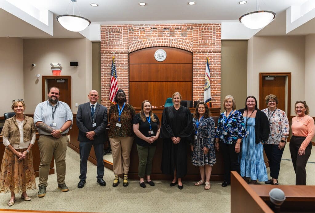 A group of eleven people, including a judge in a black robe, pose together for a photo in a courtroom with a brick judge's bench and flags behind them.